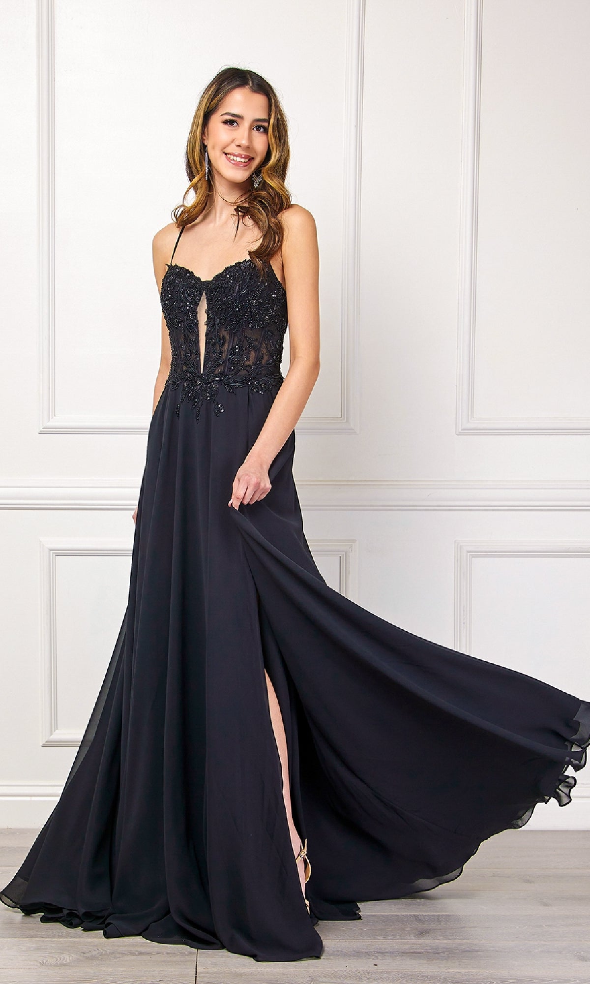 A woman stands elegantly against a white paneled wall, wearing the Long Black Corset Prom Dress (Amelia Couture 7066), featuring a sleek silhouette and flowing skirt.